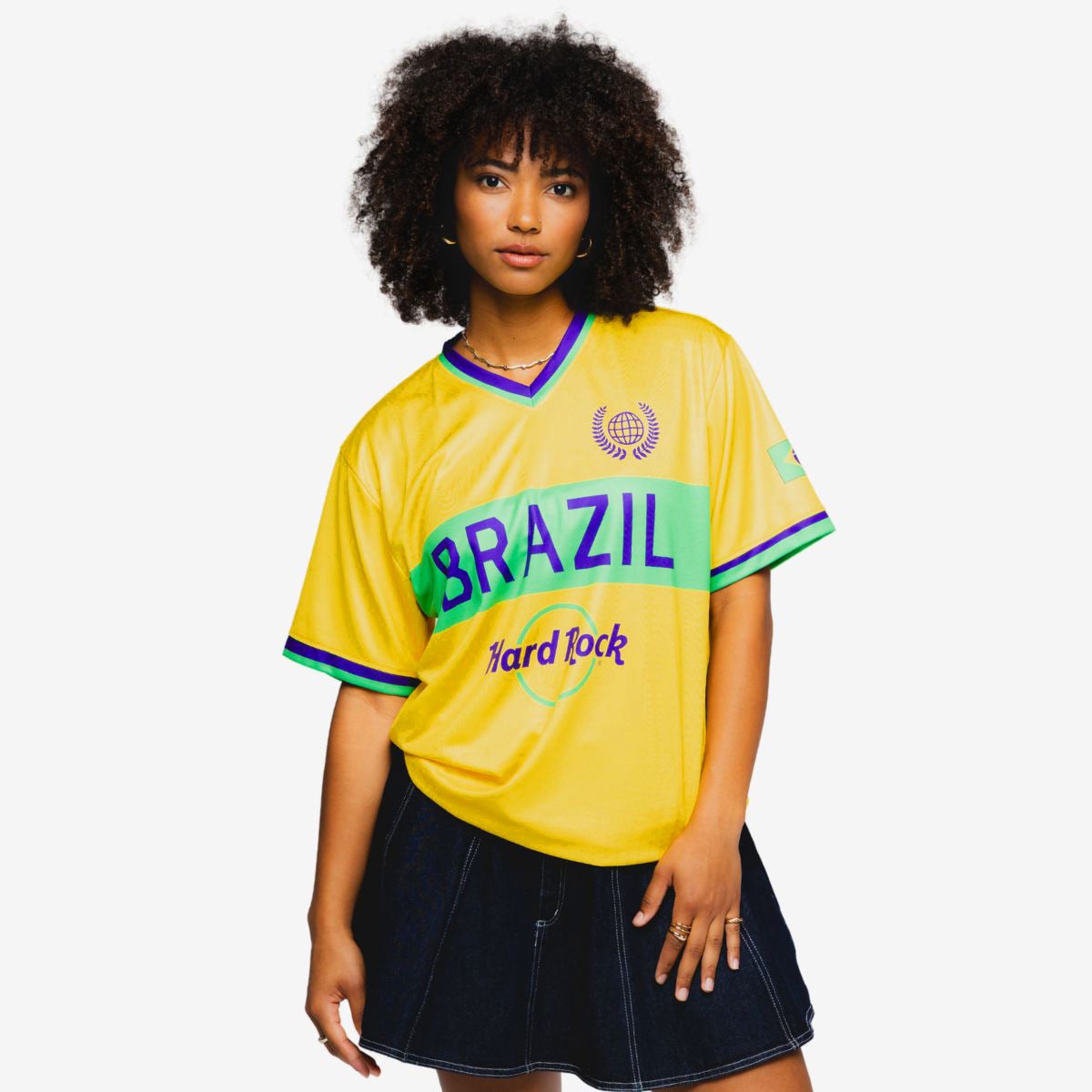 A young woman stands against a plain white background wearing a bright yellow Brazil soccer jersey. The shirt features green and blue accents with the word "BRAZIL" prominently displayed across the chest. Additional text reading "Hard Rock" appears below the main lettering. She pairs the jersey with a dark denim skirt and has one hand resting on her hip, creating a casual, sporty look.