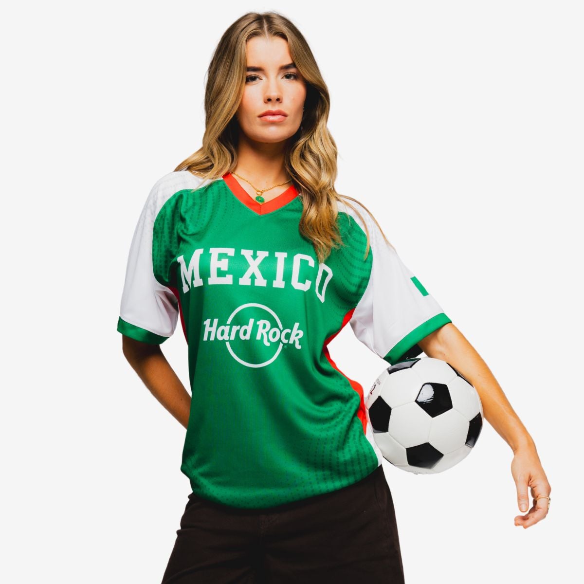A woman wearing a green Mexico soccer jersey stands against a plain white background. The shirt features the words "MEXICO" and "Hard Rock" on the front. She rests one hand on her hip while the other holds a classic black and white soccer ball at her side. The overall scene emphasizes team spirit and casual sports fashion.