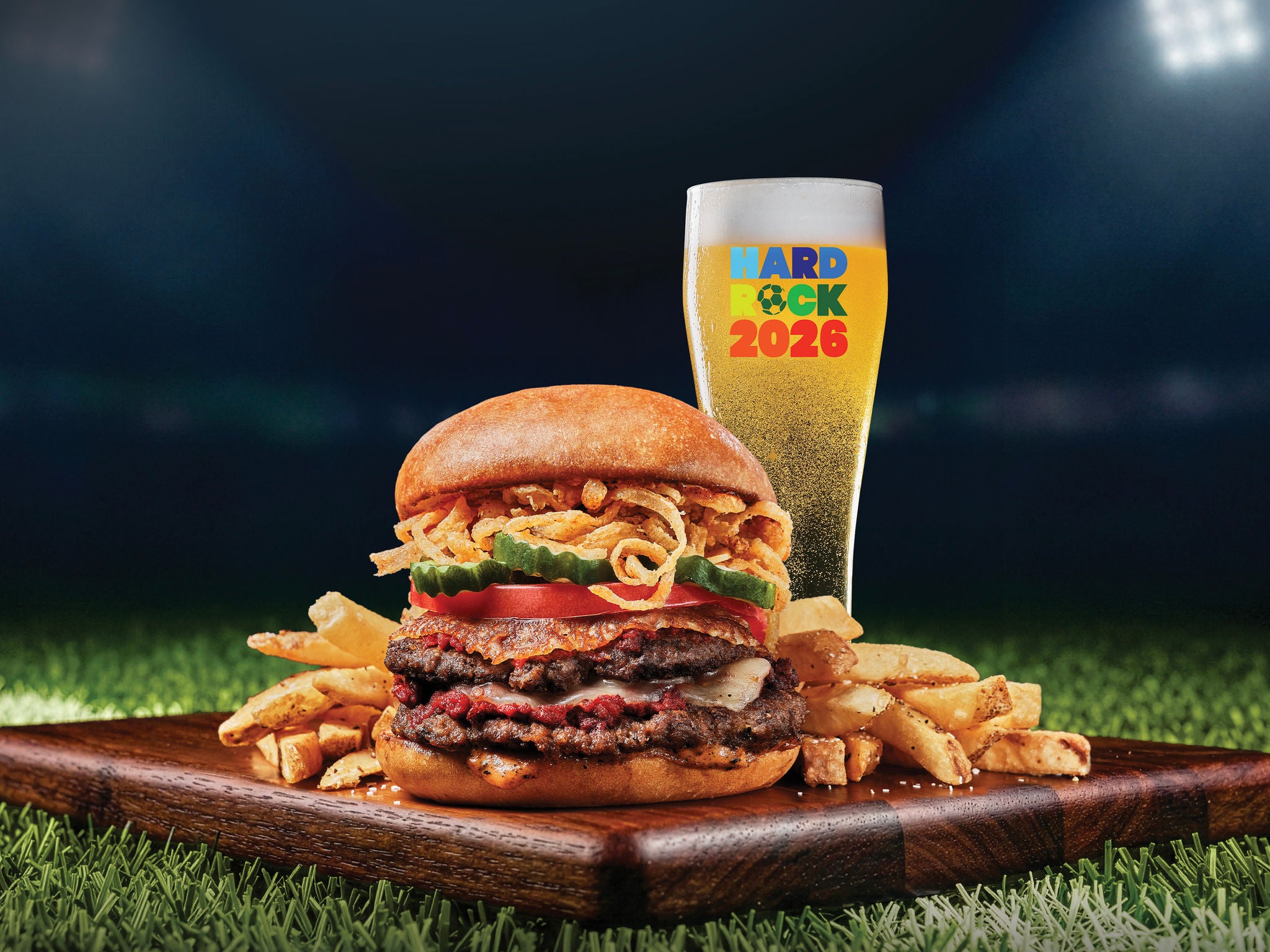 A stacked cheeseburger with multiple beef patties, fried onions, pickles, and tomato sits on a wooden board surrounded by golden French fries. Beside it stands a tall glassed drink with a foamy head, placed on a surface resembling a grass sports field. The background is dark and blurred, emphasizing the food and drink in the foreground. The glass features colorful text reading HARD ROCK 2026.